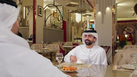 صداقة وتعارف وبناء علاقات اجتماعية, مقهى ومطعم راقي، مشاركة المشروب الساخن والحلوى، قضاء وقت ممتع في الخارج، جلسة وناسة في عطلة نهاية الاسبوع, رجل عربي خليجي اماراتي يرتدي الكندورة و الغتره يجلس مع صديقه في مطعم يتناولان القهوه