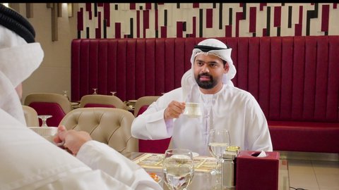 مقهى ومطعم راقي، مشاركة المشروب الساخن والحلوى، قضاء وقت ممتع في الخارج، صداقة وتعارف وبناء علاقات اجتماعية، جلسة وناسة في عطلة نهاية الاسبوع, رجل عربي خليجي اماراتي يرتدي الكندورة و الغتره يجلس مع صديقه في كوفي شوب يتناولان القهوه
