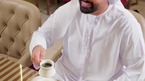 الاستمتاع باحتساء المشروب المفضل،تصوير مقرب لرجل عربي خليجي اماراتي يرتدي الكندورة و الغتره يجلس داخل المطعم يمسك بيديه كوب من القهوة, ايماءات السعادة و السرور ، تعديل المزاج بشرب الكافيين, قضاء وقت ممتع فى الخارج