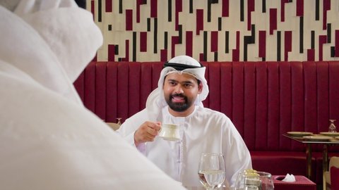 جلسة وناسة وتعارف، قضاء وقت ممتع في الخارج، شابان عربيان خليجيان اماراتيان يرتديان الكندورة و الغتره يجلسان في إحدى المطاعام الراقية بايماءات السعادة والسرور، تبادل الاحاديث بين الأصدقاء، مشاركة المشروبات والحلويات، تطوير العلاقات الاجتماعية