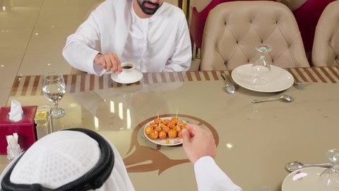 Sharing drinks and sweets, having a good time outdoors, two Emirati Gulf Arab friends wearing kandura and ghutrah sitting in a fine restaurant with gestures of happiness and joy, exchanging conversations between friends.