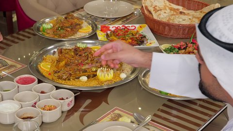 مائدة طعام مليئة بأصناف من المأكولات والمقبلات الشهية، تناول الطعام في المطاعم الإماراتية، تصوير مقرب من الخلف رجل عربي خليجي اماراتي يرتدي الكندورة يجلس على الطاولة يقوم بسكب التوابل على الطعام ، أكلات شعبية عربية خليجية اماراتية