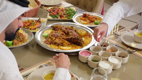 تناول الطعام في المطاعم الإماراتية، تصوير مقرب ليد رجل عربي خليجي اماراتي يرتدي الكندورة يجلس على  الطاولة يقوم بسكب التوابل على الطعام ، أكلات شعبية عربية خليجية اماراتية، مائدة طعام مليئة بأصناف من المأكولات والمقبلات الشهية