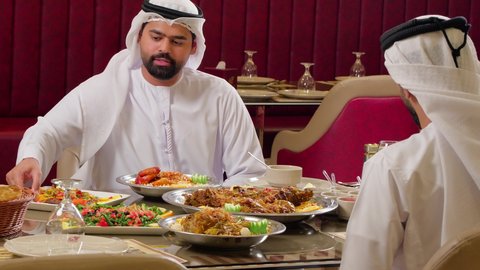 A gathering of friends in a luxurious restaurant in the UAE, serving popular Arab Gulf Emirati dishes, delicious meals, an Arab Gulf Emirati man wearing the kandura and ghutrah presenting a plate of food to his friend, gestures of happiness and joy.
