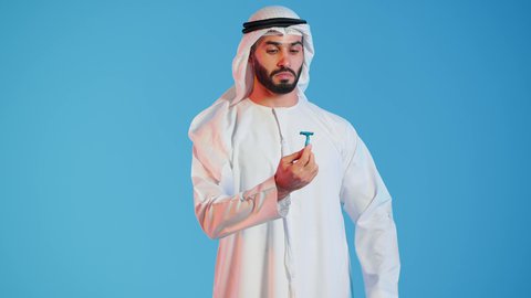 شفرة حلاقة رجالية، مفهوم العناية بالبشرة والنظافة ، بورتريه لرجل عربي خليجي اماراتي يرتدي الكندورة والغترة البيضاء يقوم بإزالة شعر الوجه بإستخدام شفرة الحلاقة ، الإهتمام المظهر الخارجي ، خلفية ملونة