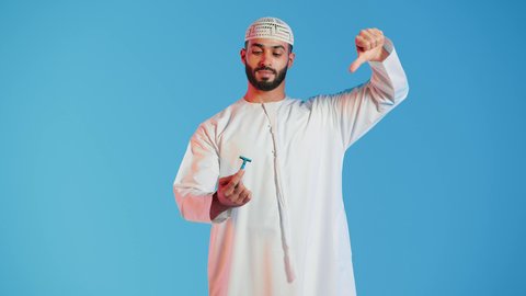 الإهتمام المظهر الخارجي ، بورتريه  لرجل عربي خليجي اماراتي يرتدي الزي الاماراتي يمسك بيده شفرة الحلاقة بايماءات عدم الاعجاب ، مفهوم العناية بالبشرة والنظافة ، شفرة حلاقة رجالية، خلفية ملونة