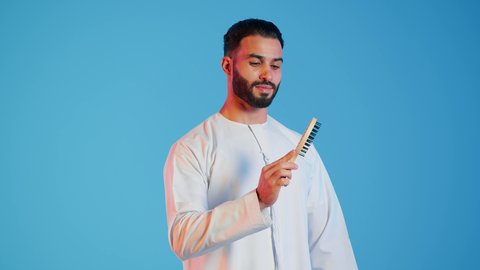 تمشيط وترتيب الشعر، مفهوم العناية والاهتمام بالجمال والأناقة والمظهر الخارجي، بورتريه لرجل عربي خليجي اماراتي يقوم بالنظر الى فرشاة الشعر ( المشط ) بتعابير وإيماءات السعادة والسرور،، خلفية ملونة