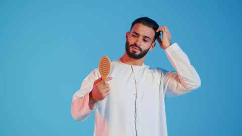 مفهوم العناية والاهتمام بالجمال والأناقة والمظهر الخارجي، تمشيط وترتيب الشعر، بورتريه لرجل عربي خليجي اماراتي يقوم بالنظر الى فرشاة الشعر ( المشط ) بتعابير وإيماءات السعادة والسرور،، خلفية ملونة