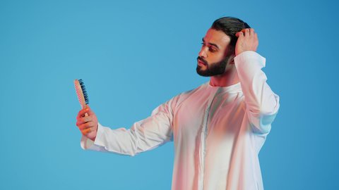 بورتريه لرجل عربي خليجي اماراتي يقوم بتصفيف شعره باستخدام فرشاة الشعر ( المشط ) بتعابير وإيماءات السعادة والسرور ،،تمشيط وترتيب الشعر، مفهوم العناية والاهتمام بالجمال والأناقة والمظهر الخارجي،  خلفية ملونة