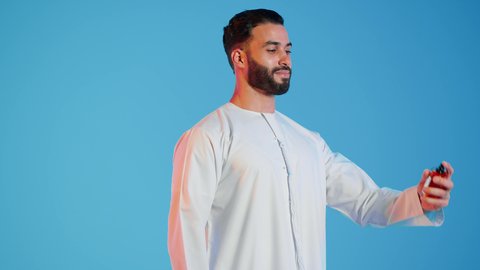 مفهوم الأناقة والإهتمام المظهر الخارجي، عطور ذات روائح فاخرة وفريدة، التسويق لمنتجات العطور، العود العطري، بورتريه مقرب لرجل عربي خليجي اماراتي يرتدي الزي الاماراتي، يقوم بوضع العطر، التطيب والتعطر  بالمناسبات والأعياد وحفلات الزفاف،  خلفيه ملونه