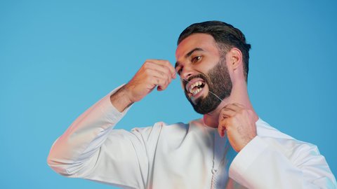 مفهوم العناية الشخصية، بورتريه مقرب لرجل عربي خليجي إماراتي مبتسم يقوم باستخدام خيط تنظيف الأسنان، مفهوم العناية والاهتمام بصحة ونظافة الفم والأسنان، روتين النظافة اليومية لتجنب تسوس الأسنان وأمراض اللثة، خلفية ملونة
