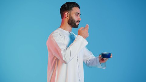 استخدام مكونات تجميلية شبابية، مفهوم العناية والإهتمام بنضارة وتحسين البشرة، ترطيب وتنظيف وتفتيح الجلد، بورتريه لرجل عربي خليجي اماراتي  يرتدي االزي  التقليدي يمسك بيده كريم المرطب ويضعه على الوجه، خلفية ملونه