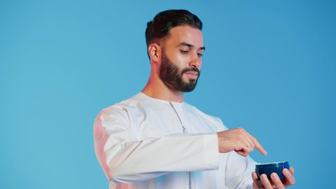 مفهوم العناية والإهتمام بنضارة وتحسين البشرة، استخدام مكونات تجميلية شبابية، ترطيب وتنظيف وتفتيح الجلد، بورتريه لرجل عربي خليجي اماراتي  يرتدي االزي  التقليدي يمسك بيده كريم المرطب ويضعه على الوجه، خلفية ملونه