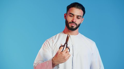 النظر الى الكاميرا بتعابير الجدية، عدم القدرة على التوقف من الكلام، الاهتمام بالمظهر الخارجي والأناقة، مفهوم السكوت والصمت، بورتريه لرجل عربي خليجي اماراتي يرتدي الزي التقليدي يحمل بيده مقص ، الوقوف باستقامة وثبات،  خلفية ملونة