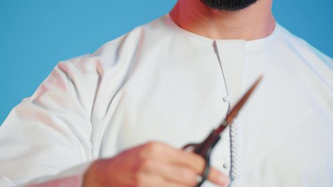 مفهوم السكوت والصمت، الاهتمام بالمظهر الخارجي والأناقة، عدم القدرة على التوقف من الكلام،  بورتريه لرجل عربي خليجي اماراتي يرتدي الزي التقليدي يحمل بيده مقص ينظر الى الكاميرا بتعابير الجدية، الوقوف باستقامة وثبات،  خلفية ملونة