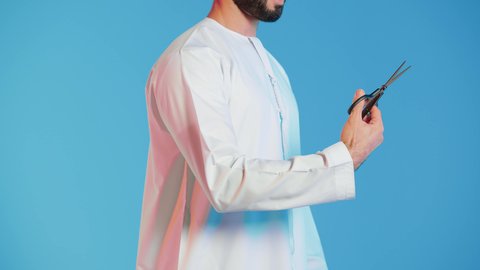 عدم القدرة على التوقف من الكلام، الاهتمام بالمظهر الخارجي والأناقة، مفهوم السكوت والصمت، بورتريه لرجل عربي خليجي اماراتي يرتدي الزي التقليدي يحمل بيده مقص ينظر الى الكاميرا بتعابير الجدية، الوقوف باستقامة وثبات،  خلفية ملونة