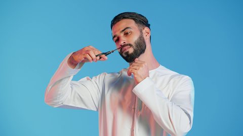 بورتريه لرجل عربي خليجي اماراتي يرتدي الثوب التقليدي يحمل بيده المقص، مركز تجميلي رجالي، مفهوم العناية و الاهتمام بالجمال والمظهر الخارجي، قص و تصفيف الذقن والشارب في صالون الحلاقة، خلفية ملونة.
