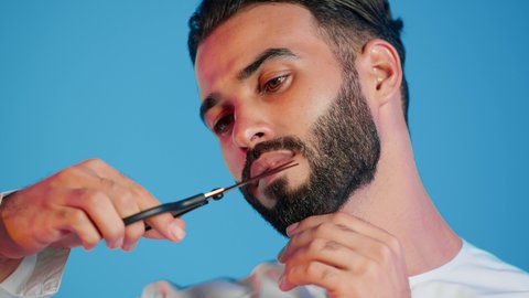 قص و تصفيف الذقن والشارب في صالون الحلاقة، مركز تجميلي رجالي، مفهوم العناية و الاهتمام بالجمال والمظهر الخارجي،بورتريه لرجل عربي خليجي اماراتي يرتدي الثوب التقليدي يحمل بيده المقص، خلفية ملونة.