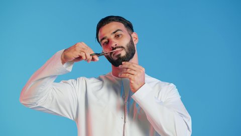 مركز تجميلي رجالي، مفهوم العناية و الاهتمام بالجمال والمظهر الخارجي، قص و تصفيف الذقن والشارب في صالون الحلاقة، بورتريه لرجل عربي خليجي اماراتي يرتدي الثوب التقليدي يحمل بيده المقص، خلفية ملونة.