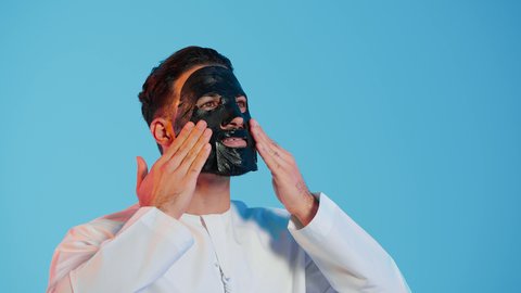 خطوات تنظيف وتقشير البشرة،استخدام مكونات تجميلية،  بورتريه لرجل عربي خليجي اماراتي يرتدي الزي التقليدي يقوم بوضع قناع الطين على وجهه، مفهوم العناية والاهتمام بالمظهر الخارجي، خلفية ملونة.