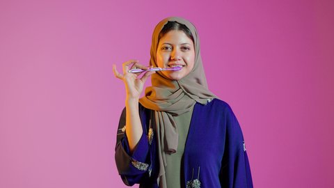 بورتريه مقرب لسيدة عربية خليجية اماراتيه تقوم بتنظيف الأسنان بفرشاة الأسنان ، مفهوم العناية والإهتمام بصحة ونظافة الأسنان ، روتين نظافة الفم اليومية ، تجنب تسوس الاسنان وامراض اللثة ، خلفية ملونة