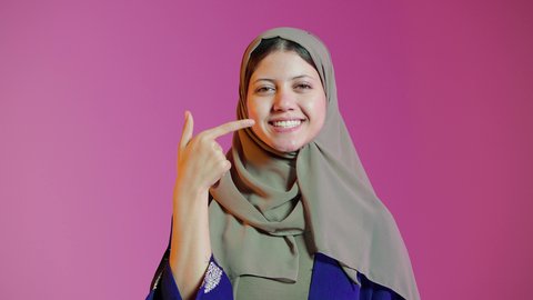 استخدام قشور فينير من الخزف للأسنان،تجميل وتبييض الاسنان، بورتريه لامراه عربيه خليجيه اماراتيه مبتسمه  وتشير بإصبعه السبابة على أسنانها ، إبتسامة هوليود، معالجة الأسنان، أسنان نظيفة مثالية ,خلفية ملونة