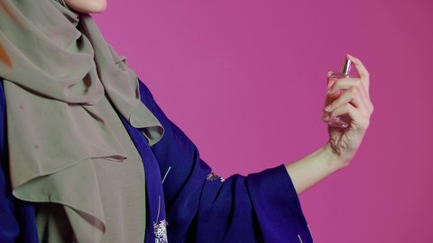 التطيب والتعطر بالعود العطري، استخدام الزيوت العطرية الطبيعية، الحيرة في اختيار الرائحة، منتجات العطور النسائيه، بورتريه لإمرأة عربية خليجية اماراتيه ترتدي العباءة والحجاب تقوم برش العطر، الأناقة والإهتمام بالمظهر الخارجي، خلفية ملونة