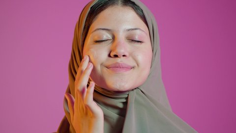 الاهتمام بالمظهر الخارجي، مفهوم العناية بنضارة وتحسين البشرة، بورتريه مقرب لامرأة عربية خليجية اماراتيه محجبة مبتسمة تضع كريم الترطيب، استخدام كريمات ومكونات تجميلية، خلفية ملونة