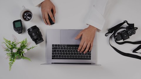 ادخال الصور على جهاز الحاسوب ، الاستمتاع بالمشروبات الساخنه ،تصوير مقرب من الأعلى لمصور سعودي عربي يعمل من خلال اللابتوب على مكتب أبيض , مهن ووظيفة سعودية , بيئة عمل مكتبية , خلفية بيضاء .