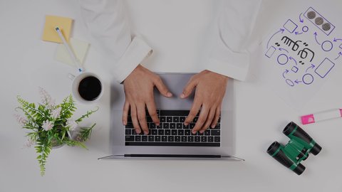 An expert marketer working on a laptop at a white desk, a close-up shot from above of a Saudi Arabian man with experience in business management, a Saudi job and profession, an office work environment, white background.
