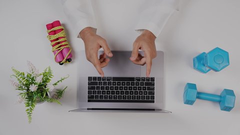A close-up shot from above of a fitness trainer working on a laptop at a white desk, focusing on physical fitness, a profession and job in Saudi Arabia, an office work environment, with a white background.
