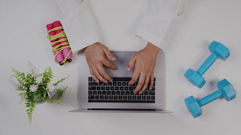 بيئة عمل مكتبية ، الاهتمام باللياقة البدنية ،تصوير  مقرب من الأعلى لمدرب رياضي يعمل من خلال اللابتوب على مكتب أبيض , مهنة ووظيفه سعودية ,خلفية بيضاء .