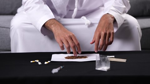 النتائج السلبية للإدمان على الصحة العامة، القيام بعادات سلوكية ضارة وتعاطي المواد السامة، الأثار السلبية للادمان على التبغ والمخدرات، تصوير مقرب لأيدي شاب عربي خليجي إماراتي يرتدي الكندورة يقوم بلف عقب السجائر