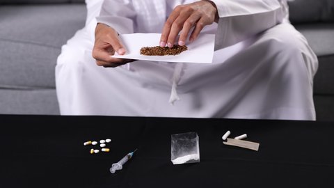 التأثير السلبي للاضطرابات النفسية على الصحة الجسدية، القيام بعادات سلوكية ضارة وتعاطي المواد السامة، الأثار السلبية للادمان على التبغ والمخدرات، تصوير مقرب لأيدي شاب عربي خليجي إماراتي يرتدي الكندورة يقوم بلف عقب السجائر