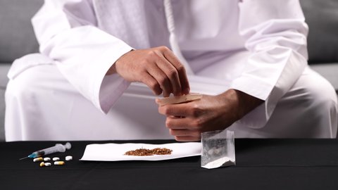 ممارسة سلوكيات ضارة، الأثار السلبية للادمان على التبغ والمخدرات، المعاناة من الاضطرابات النفسية والمشاكل الصحية، تصوير مقرب لرجل عربي خليجي إماراتي يرتدي الكندورة والغترة البيضاء يجلس على اريكة يقوم بحشو عقب السجائر