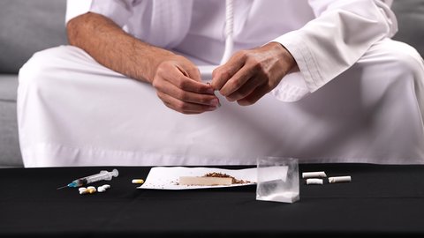 ممارسة سلوكيات ضارة واستهلاك المواد السامة، الأثار السلبية للادمان على التبغ والمخدرات، المعاناة من الاضطرابات النفسية والمشاكل الصحية، تصوير مقرب لرجل عربي خليجي إماراتي يرتدي الكندورة يقوم بحشو عقب السجائر