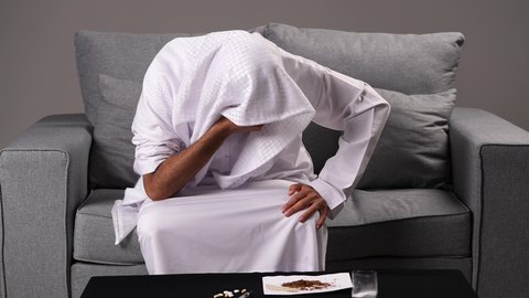 الصراع الداخلي الناتج عن تأنيب الضمير، عادات سلوكية ضارة وتعاطي مواد سامة، المعاناة من الاضطرابات النفسية والمشاكل الصحية، الادمان على التبغ والمخدرات، رجل عربي خليجي إماراتي يرتدي الكندورة يجلس أمام طاولة تحوي مجموعة من مواد المعاقرة