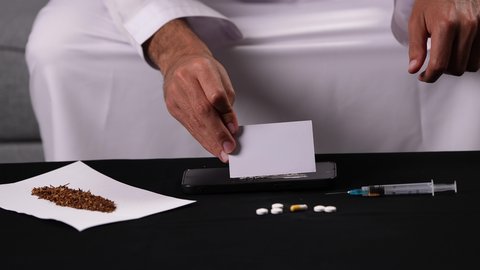 ممارسة سلوكيات ضارة، الآثار السلبية للادمان، تعاطي المواد السامة، الاضطرابات النفسية والمشاكل الصحية، تصوير مقرب لأيدي شاب عربي خليجي إماراتي يرتدي الكندورة يقوم بتحضير البودرة البيضاء لاستنشاقها