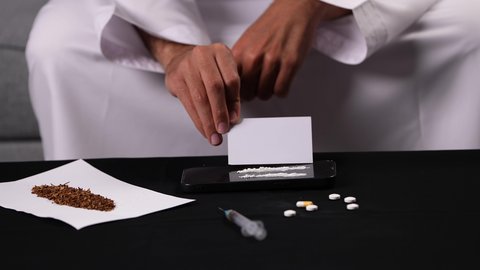 القيام بعادات سلوكية ضارة، الآثار السلبية للادمان، تعاطي المواد السامة، الاضطرابات النفسية والمشاكل الصحية، تصوير مقرب لأيدي شاب عربي خليجي إماراتي يرتدي الكندورة يقوم بتحضير البودرة البيضاء لاستنشاقها