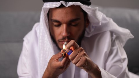 Suffering from psychological disorders and health problems, engaging in harmful behavioral habits and substance abuse, the negative effects of addiction to tobacco and drugs, a close-up shot of an Emirati Gulf Arab man wearing a kandura and ghutrah smoking hashish cigarettes.
