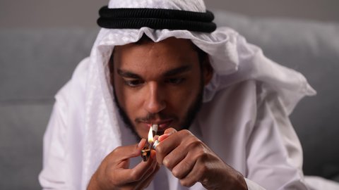 القيام بعادات سلوكية ضارة وتعاطي المواد السامة، الأثار السلبية للادمان على التبغ والمخدرات، المعاناة من الاضطرابات النفسية والمشاكل الصحية، تصوير مقرب لرجل عربي خليجي اماراتى يرتدي الكندورة و الغترة يقوم بتدخين سجائر الحشيش المخدر