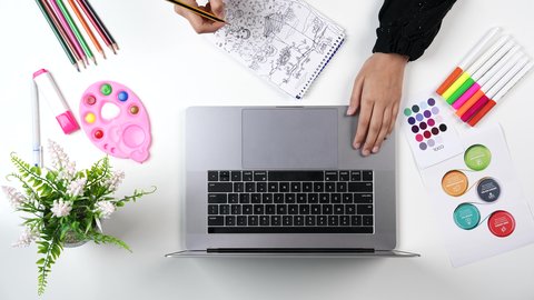 A close-up shot from above of the hand of a Saudi Arabian Gulf woman drawing on a canvas in front of a laptop, learning and creativity, skill development, coloring tools and equipment, artistic recreational activities.