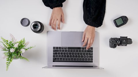 نقل الصور إلى جهاز الحاسوب، تصوير مقرب من الأعلى لمصورة سعودية عربية تعمل من خلال اللابتوب على مكتب أبيض , مهن ووظيفة سعودية , بيئة عمل مكتبية , خلفية بيضاء