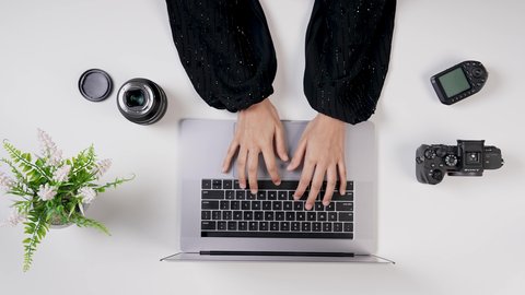 ادخال الصور على جهاز الحاسوب  ،تصوير مقرب من الأعلى لمصورة سعودية عربية تعمل من خلال اللابتوب على مكتب أبيض , مهن ووظيفة سعودية , بيئة عمل مكتبية , خلفية بيضاء