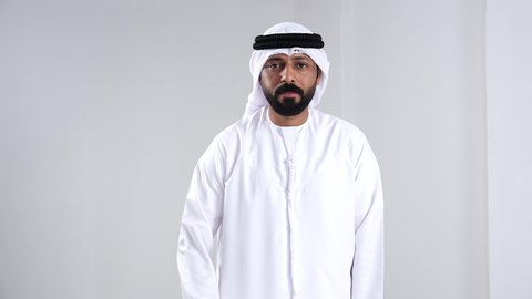 Gestures of happiness and joy, the command to stop doing something, expressions of agreement and acceptance, a portrait of an Emirati Gulf Arab man wearing a kandura and ghutrah looking at the camera raising his thumb up, white background.