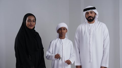 امتلاك منزل خاص, الالتزام بالقواعد والقوانين الأسريه، بورتريه لولد عربي خليجي اماراتي يرتدي الكندورة و الغتره يحمل مفتاح المنزل الجديد برفقة والدية، ايماءات تدل على السعادة والفرح ، أساسيات العائلة الاماراتية السعيده، خلفية بيضاء