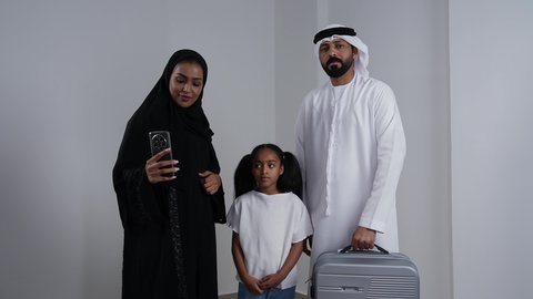 Documenting happy moments through mobile phones, using modern devices and technologies, a portrait of an Emirati Gulf Arab family wearing traditional attire taking commemorative selfies with gestures of joy and happiness, an Emirati Gulf Arab man wearing a kandura and ghutrah holding a travel bag in preparation for a recreational trip, white background.