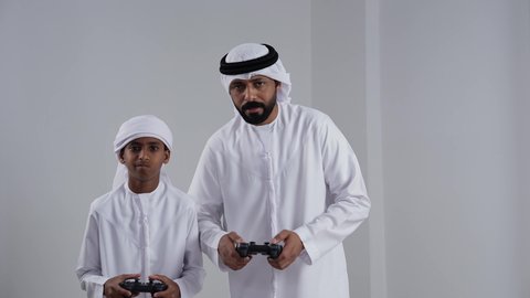 ايماءات وجه ويدين تدل الاندماج و التركيز, الاستمتاع بلعب العاب الفيديو, مفهوم الفوز والخسارة, بورتريه لرجل عربي خليجي اماراتي يرتدي الكندورة و الغتره يمسك عصا التحكم برفقة ابنه، خلفية بيضاء