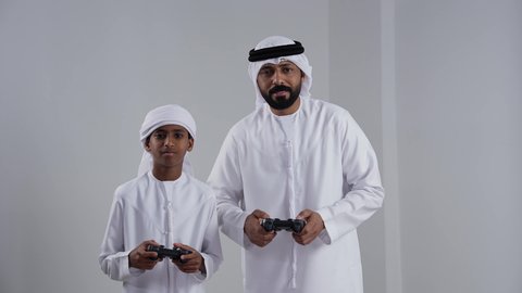 الاستمتاع بلعب العاب الفيديو, مفهوم الفوز والخسارة, بورتريه لرجل عربي خليجي اماراتي يرتدي الكندورة و الغتره يمسك عصا التحكم برفقة ابنه، ايماءات وجه ويدين تدل على الفوز والانتصار، خلفية بيضاء