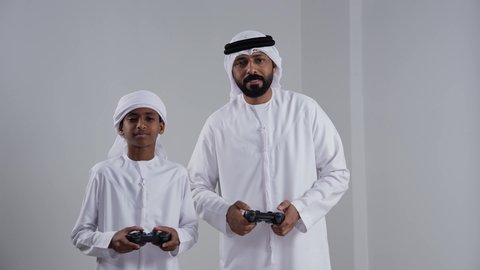 مفهوم الفوز والخسارة, بورتريه لرجل عربي خليجي اماراتي يرتدي الكندورة و الغتره يمسك عصا التحكم برفقة ابنه، الاستمتاع بلعب العاب الفيديو، ايماءات وجه ويدين تدل على الفوز والانتصار، خلفية بيضاء